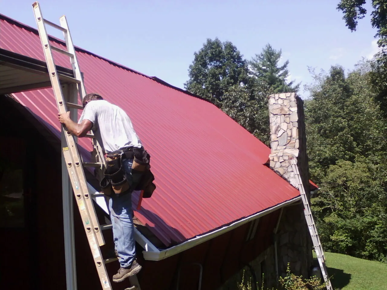 Roofer on a ladder beside a red metal roof and stone chimney — Soffit & Fascia Repair by Roofing Brothers in Kingsbury